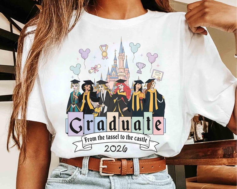 May include: White t-shirt featuring a colorful graphic of Disney princesses in graduation attire in front of a castle. The shirt includes the text "Graduate" and "From the tassel to the castle 2026".
