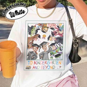 May include: White t-shirt with a colorful graphic featuring characters from the Toy Story franchise wearing graduation caps. The text "TO KINDERGARTEN AND BEYOND" is below the graphic. The shirt also has the words "COMFORT COLORS" and "White" in a white oval.