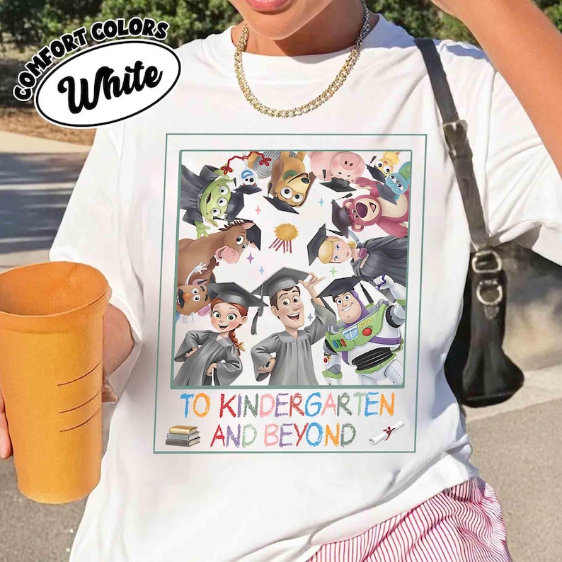 May include: White t-shirt with a colorful graphic featuring characters from the Toy Story franchise wearing graduation caps. The text "TO KINDERGARTEN AND BEYOND" is below the graphic. The shirt also has the words "COMFORT COLORS" and "White" in a white oval.