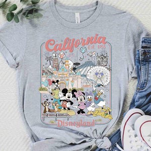 Puede incluir: Camiseta gris con un gr&aacute;fico colorido de Disneyland en California. El dise&ntilde;o incluye a Mickey y Minnie Mouse, y el texto "California Disneyland Est. 1955". La camiseta se combina con vaqueros azules y zapatillas blancas.