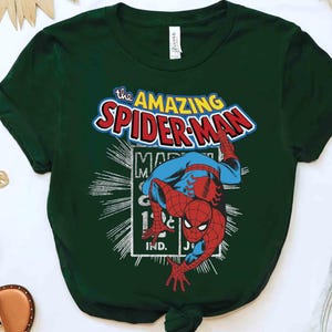 May include: Dark green t-shirt with a vintage Spider-Man graphic. The design features the words "The Amazing Spider-Man" in red, blue, and white, and an illustration of Spider-Man. The shirt is made of a soft material and is suitable for casual wear.