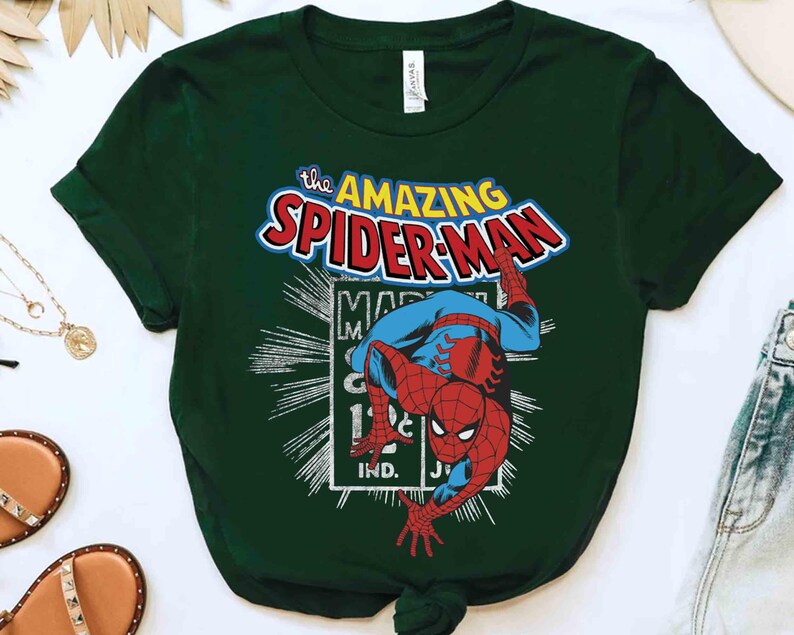 May include: Dark green t-shirt with a vintage Spider-Man graphic. The design features the words "The Amazing Spider-Man" in red, blue, and white, and an illustration of Spider-Man. The shirt is made of a soft material and is suitable for casual wear.