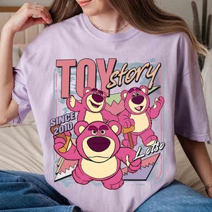 May include: A lilac t-shirt with a graphic of Lotso, a pink bear character from the film 'Toy Story'. The design includes the text 'TOY story' and 'SINCE 2010'.