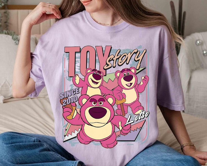May include: A lilac t-shirt with a graphic of Lotso, a pink bear character from the film 'Toy Story'. The design includes the text 'TOY story' and 'SINCE 2010'.