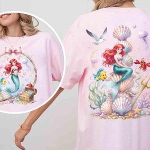 May include: A light pink t-shirt featuring a large graphic print of a red-haired mermaid sitting on a stack of seashells, surrounded by a yellow fish, a red crab, a seagull, a golden trident, and various colorful coral and sea life.