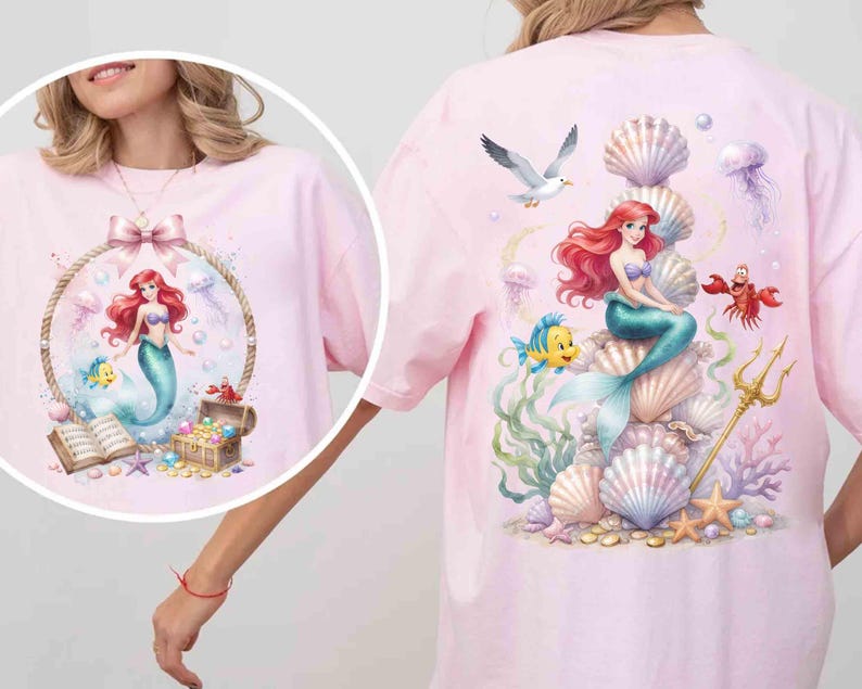 May include: A light pink t-shirt featuring a large graphic print of a red-haired mermaid sitting on a stack of seashells, surrounded by a yellow fish, a red crab, a seagull, a golden trident, and various colorful coral and sea life.