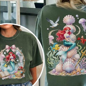 May include: A dark green t-shirt featuring a graphic of a red-haired mermaid with a blue tail, a yellow fish, a crab, and a treasure chest. The back design shows the mermaid sitting on a stack of seashells with a trident, jellyfish, and a seagull.