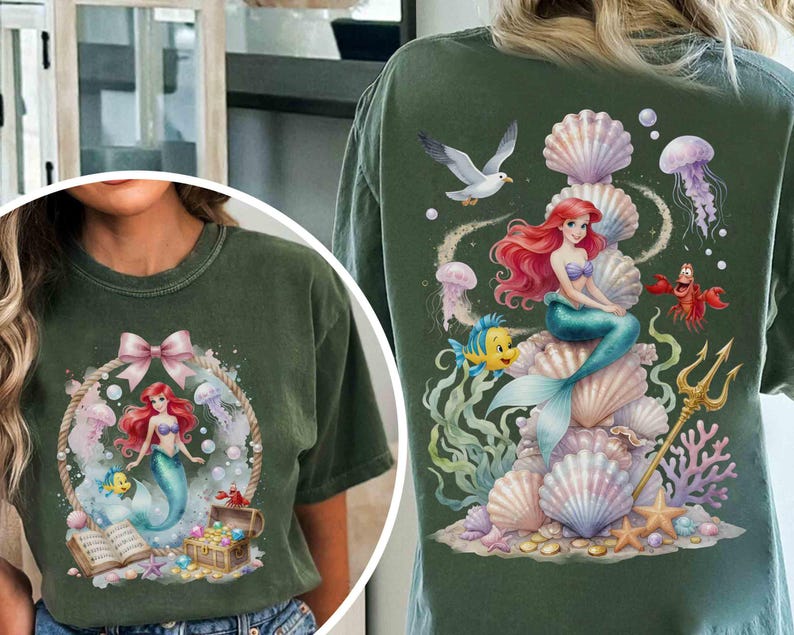 May include: A dark green t-shirt featuring a graphic of a red-haired mermaid with a blue tail, a yellow fish, a crab, and a treasure chest. The back design shows the mermaid sitting on a stack of seashells with a trident, jellyfish, and a seagull.