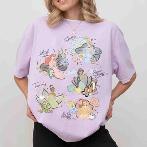 May include: A light purple t-shirt featuring a graphic print of various animated characters including Ariel, Cinderella, Jasmine, Tiana, and Belle. Each character is accompanied by their name in cursive script, small icons, and scattered star accents.