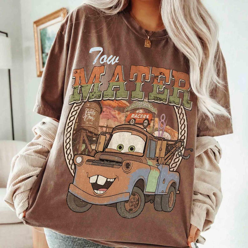 K&ouml;nnte beinhalten: Ein braunes Kurzarm-T-Shirt mit einer Grafik der Figur Tow Mater aus dem Film Cars. Das Design enth&auml;lt den Text Tow Mater, einen Wasserturm und ein kleines orangefarbenes Auto mit der Aufschrift Racers. Die Grafik ist von einem Seilkreis umrahmt.