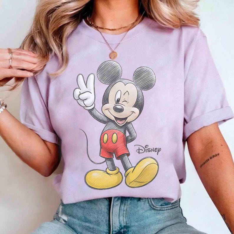 May include: A light purple t-shirt featuring a hand-drawn image of Mickey Mouse giving a peace sign. Mickey is wearing red shorts, yellow shoes, and black gloves. The Disney logo is printed below the image.