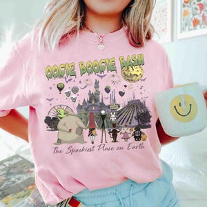 Puede incluir: Una camiseta rosa claro con un estampado gr&aacute;fico que incluye el texto OOGIE BOOGIE BASH y The Spookiest Place on Earth. El dise&ntilde;o muestra personajes de Pesadilla antes de Navidad, un castillo, una luna, fuegos artificiales y murci&eacute;lagos.