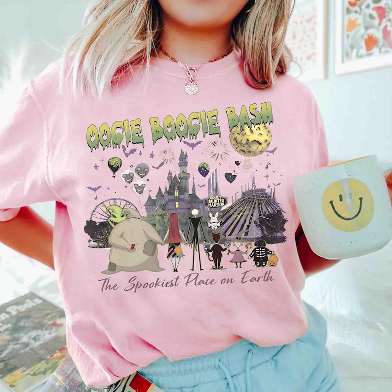 Puede incluir: Una camiseta rosa claro con un estampado gr&aacute;fico que incluye el texto OOGIE BOOGIE BASH y The Spookiest Place on Earth. El dise&ntilde;o muestra personajes de Pesadilla antes de Navidad, un castillo, una luna, fuegos artificiales y murci&eacute;lagos.