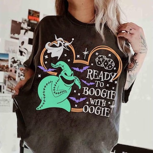 以下が含まれることがあります： 緑色のウギー・ブギーのキャラクターデザインと「Ready to Boogie with Oogie」の文字が入った濃いグレーのTシャツ。デザインにはコウモリ、サイコロ、幽霊が含まれています。Tシャツはクルーネックで半袖です。