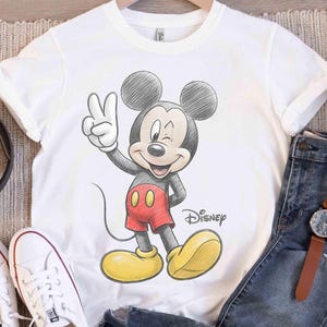 May include: White t-shirt featuring a sketch-style illustration of Mickey Mouse winking and giving a peace sign. Mickey is wearing red shorts, yellow shoes, and black gloves. The Disney logo is printed below the character.