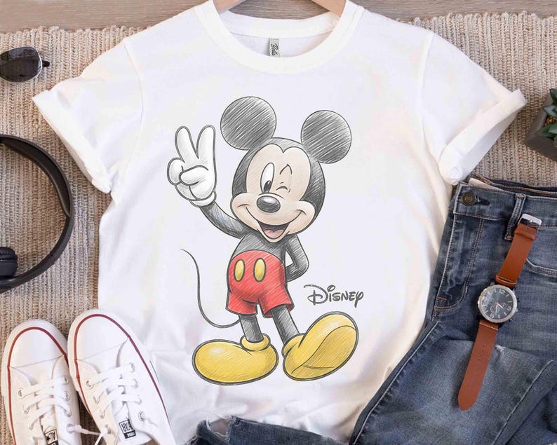 May include: White t-shirt featuring a sketch-style illustration of Mickey Mouse winking and giving a peace sign. Mickey is wearing red shorts, yellow shoes, and black gloves. The Disney logo is printed below the character.
