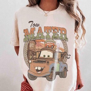 K&ouml;nnte beinhalten: Ein cremefarbenes T-Shirt mit einem Grafikdruck der Figur Tow Mater. Das Design enth&auml;lt den Text Tow Mater, eine W&uuml;stenlandschaft, einen Wasserturm und ein kleines Rennauto. Die Grafik verwendet ged&auml;mpfte Orange-, Gr&uuml;n- und Braunt&ouml;ne.