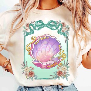 Puede incluir: Camiseta color crema con un dise&ntilde;o de concha. La concha est&aacute; representada en tonos morados y rosas, con detalles dorados. El dise&ntilde;o est&aacute; enmarcado por un borde turquesa, un lazo turquesa y detalles florales. Es una prenda de vestir informal.