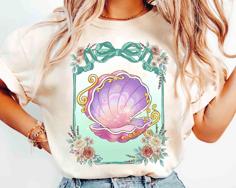 Puede incluir: Camiseta color crema con un dise&ntilde;o de concha. La concha est&aacute; representada en tonos morados y rosas, con detalles dorados. El dise&ntilde;o est&aacute; enmarcado por un borde turquesa, un lazo turquesa y detalles florales. Es una prenda de vestir informal.
