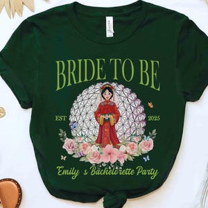 K&ouml;nnte beinhalten: Dunkelgr&uuml;nes T-Shirt mit dem Text "BRIDE TO BE" in gr&uuml;ner Schrift. Das Shirt zeigt eine Cartoon-Braut in einem roten Kleid, umgeben von Blumen und Schmetterlingen. Der Text "EST 2025" und "Emily's Bachelorette Party" sind ebenfalls sichtbar.