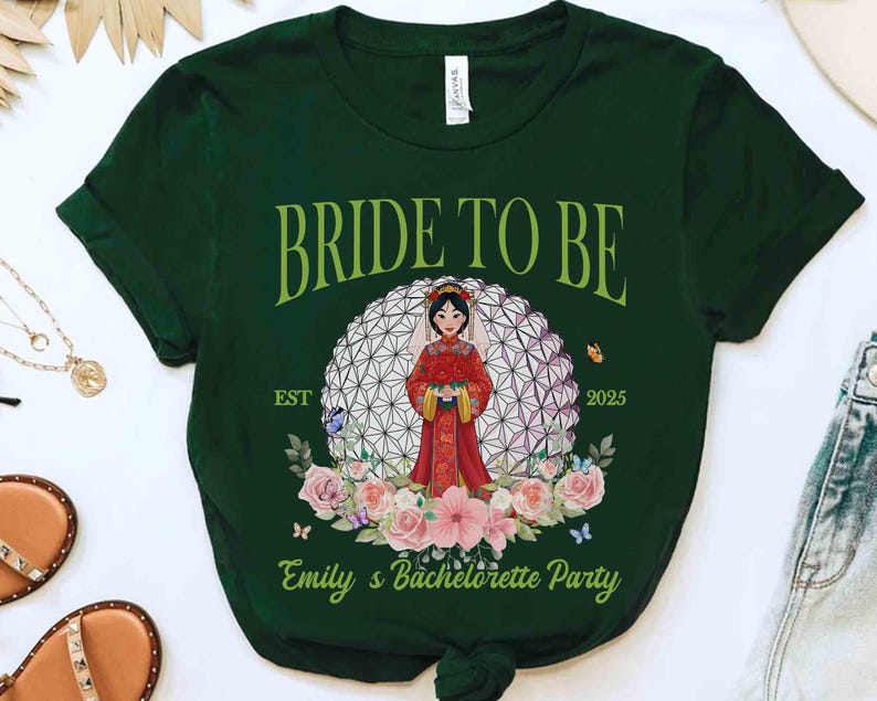 K&ouml;nnte beinhalten: Dunkelgr&uuml;nes T-Shirt mit dem Text "BRIDE TO BE" in gr&uuml;ner Schrift. Das Shirt zeigt eine Cartoon-Braut in einem roten Kleid, umgeben von Blumen und Schmetterlingen. Der Text "EST 2025" und "Emily's Bachelorette Party" sind ebenfalls sichtbar.