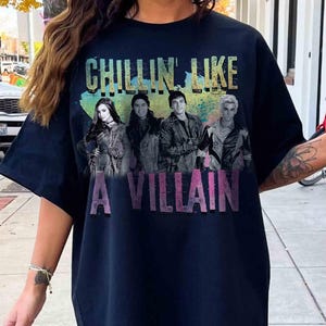 May include: Navy blue t-shirt with the text "CHILLIN' LIKE A VILLAIN" in a colorful, distressed font. The shirt features a graphic of four people. The overall design has a vintage, faded look.