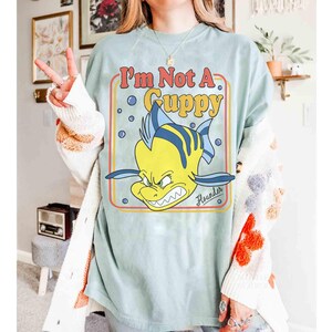 Op de afbeelding: Lichtblauw T-shirt met een cartoonafbeelding van Botje uit De Kleine Zeemeermin met de tekst "I'm Not A Guppy". De afbeelding heeft een retro-stijl met een gele, oranje en rode rand. Het woord "Flounder" staat eronder.