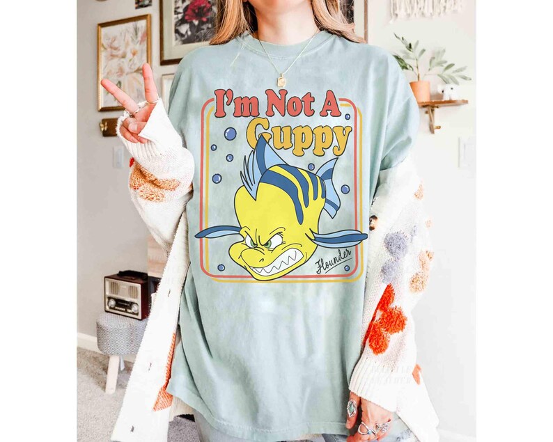 Op de afbeelding: Lichtblauw T-shirt met een cartoonafbeelding van Botje uit De Kleine Zeemeermin met de tekst "I'm Not A Guppy". De afbeelding heeft een retro-stijl met een gele, oranje en rode rand. Het woord "Flounder" staat eronder.
