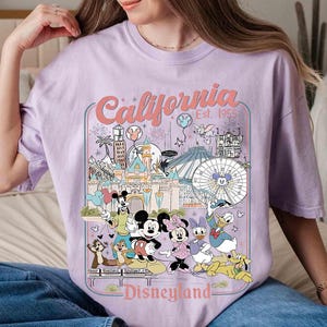 Puede incluir: Camiseta lila claro con un dise&ntilde;o gr&aacute;fico colorido de Disneyland en California. El dise&ntilde;o incluye a Mickey Mouse, Minnie Mouse, Donald Duck y otros personajes, con las palabras "California Disneyland Est. 1955".
