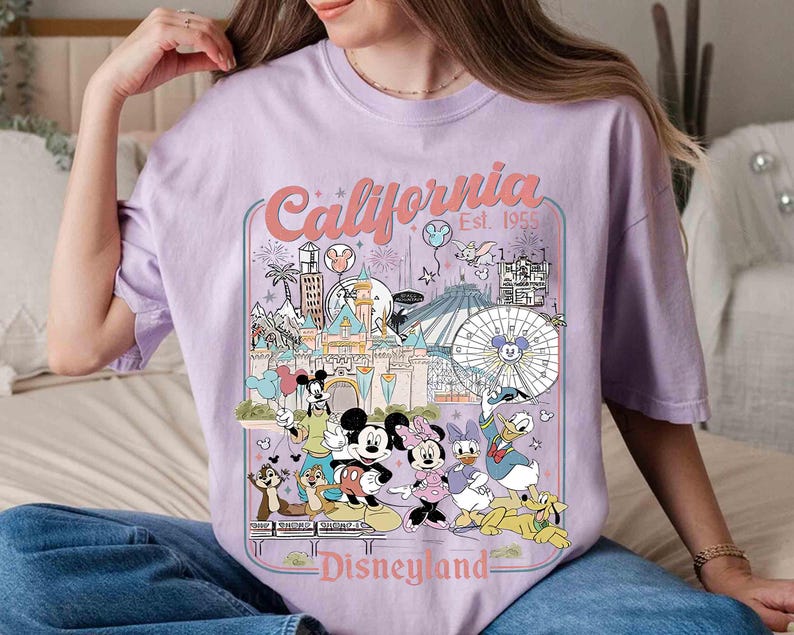 Puede incluir: Camiseta lila claro con un dise&ntilde;o gr&aacute;fico colorido de Disneyland en California. El dise&ntilde;o incluye a Mickey Mouse, Minnie Mouse, Donald Duck y otros personajes, con las palabras "California Disneyland Est. 1955".