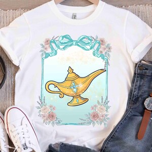 Puede incluir: Camiseta blanca con un dise&ntilde;o de l&aacute;mpara de genio dorada. La l&aacute;mpara est&aacute; enmarcada por un borde turquesa, un lazo turquesa y elementos florales. El dise&ntilde;o es de estilo caricaturesco, probablemente una prenda tem&aacute;tica.