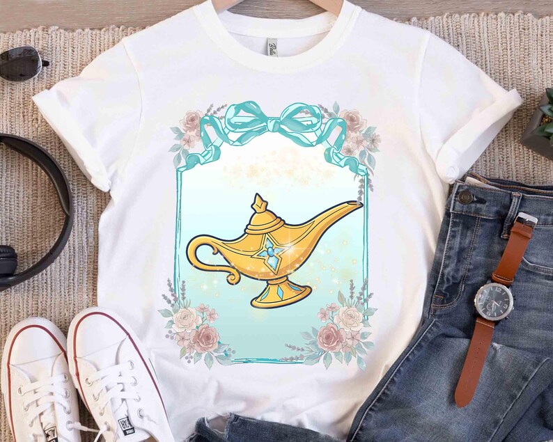 Puede incluir: Camiseta blanca con un dise&ntilde;o de l&aacute;mpara de genio dorada. La l&aacute;mpara est&aacute; enmarcada por un borde turquesa, un lazo turquesa y elementos florales. El dise&ntilde;o es de estilo caricaturesco, probablemente una prenda tem&aacute;tica.