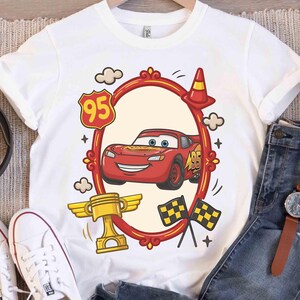 May include: White t-shirt featuring a red cartoon race car design within a red oval frame. The design includes a gold trophy, checkered flags, and the number 95. The shirt is paired with blue jeans and white sneakers.