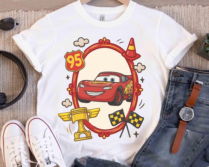 May include: White t-shirt featuring a red cartoon race car design within a red oval frame. The design includes a gold trophy, checkered flags, and the number 95. The shirt is paired with blue jeans and white sneakers.