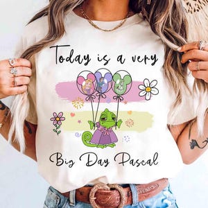 May include: Cream-colored t-shirt with the text "Today is a very Big Day Pascal." The shirt features a cartoon Pascal character holding balloons with Disney characters. Pink, purple, and yellow accents and floral designs complete the design.