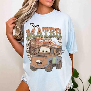 K&ouml;nnte beinhalten: Ein hellblaues T-Shirt mit einem Grafikdruck der Figur Tow Mater. Das Design enth&auml;lt den Text Tow Mater, einen Wasserturm und ein Radiator Springs Schild. Die Illustration zeigt einen rostbraunen Abschleppwagen mit gr&uuml;nen Akzenten.
