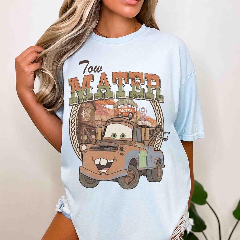 K&ouml;nnte beinhalten: Ein hellblaues T-Shirt mit einem Grafikdruck der Figur Tow Mater. Das Design enth&auml;lt den Text Tow Mater, einen Wasserturm und ein Radiator Springs Schild. Die Illustration zeigt einen rostbraunen Abschleppwagen mit gr&uuml;nen Akzenten.