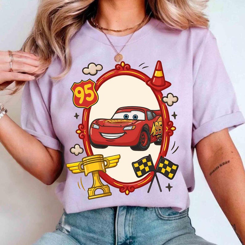 May include: A light purple t-shirt featuring a cartoon image of Lightning McQueen from the movie Cars. The design includes a red race car, a trophy, checkered flags, and the number 95.