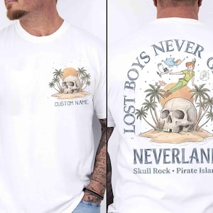 May include: White t-shirt featuring a skull on a sandy island with palm trees and a sunset. The front has "CUSTOM NAME" text. The back displays Peter Pan, the text "LOST BOYS NEVER GROW UP" and "NEVERLAND Skull Rock Pirate Island".