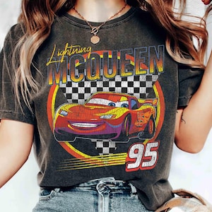 May include: Dark grey t-shirt with a graphic of Lightning McQueen from the Cars film. The design features the text "Lightning McQueen" in yellow and orange, a chequered flag, and the number "95" in white and red.