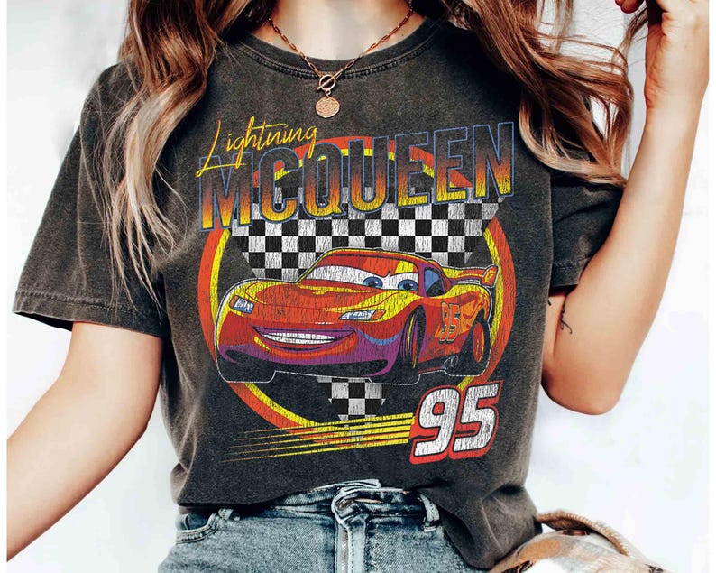 May include: Dark grey t-shirt with a graphic of Lightning McQueen from the Cars film. The design features the text "Lightning McQueen" in yellow and orange, a chequered flag, and the number "95" in white and red.