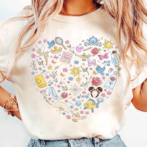 May include: Cream-colored t-shirt featuring a heart-shaped design filled with colorful illustrations of fairy tale elements. Includes a crown, rose, magic lamp, and other whimsical icons. The shirt is a comfortable, casual style.