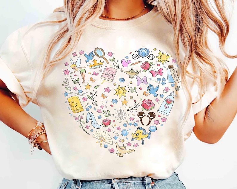 May include: Cream-colored t-shirt featuring a heart-shaped design filled with colorful illustrations of fairy tale elements. Includes a crown, rose, magic lamp, and other whimsical icons. The shirt is a comfortable, casual style.