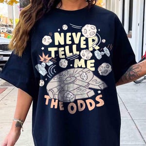 Puede incluir: Camiseta negra con un diseño gráfico que presenta el Halcón Milenario, planetas y naves espaciales. El texto "NEVER TELL ME THE ODDS" está impreso en amarillo claro y melocotón.