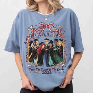 Pode incluir: Uma camiseta azul clara com um gr&aacute;fico de princesas da Disney em trajes de formatura. A palavra "GRADUATE" est&aacute; acima das princesas, com o texto "From The Tassel To The Castle 2026" abaixo.