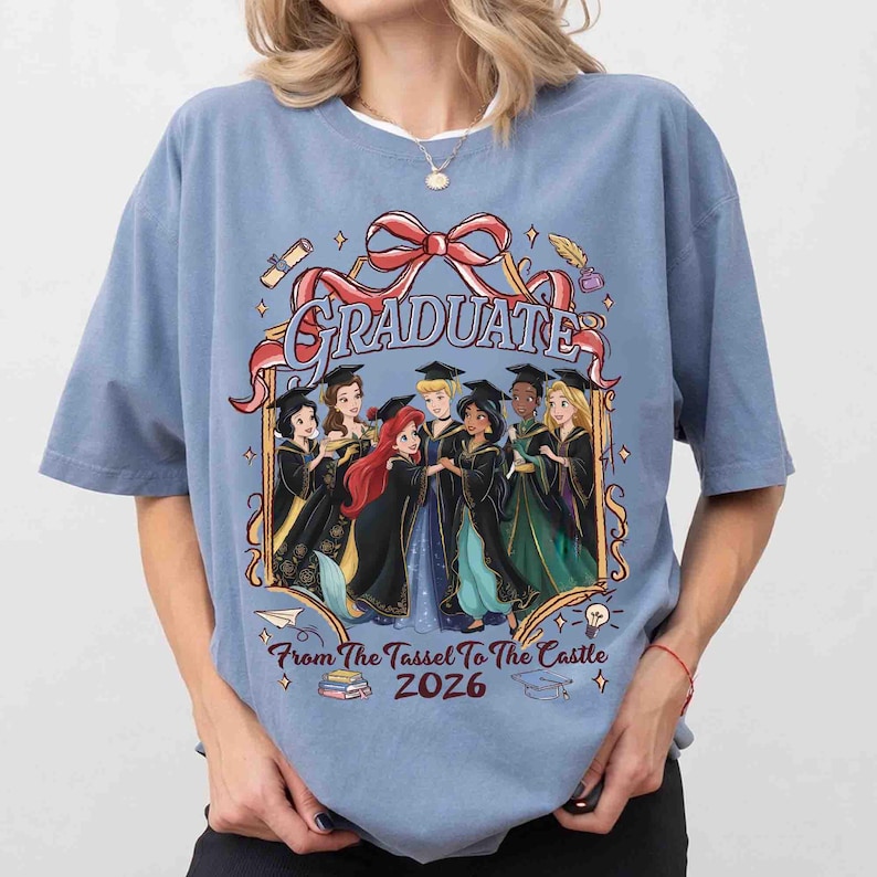 Pode incluir: Uma camiseta azul clara com um gr&aacute;fico de princesas da Disney em trajes de formatura. A palavra "GRADUATE" est&aacute; acima das princesas, com o texto "From The Tassel To The Castle 2026" abaixo.