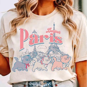 May include: Cream-colored t-shirt featuring the word "Paris" in pink with a blue outline, and the word "Bonjour" in pink. The Eiffel Tower and three cartoon kittens are also depicted. A vintage-style design.