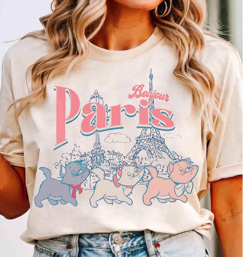 May include: Cream-colored t-shirt featuring the word "Paris" in pink with a blue outline, and the word "Bonjour" in pink. The Eiffel Tower and three cartoon kittens are also depicted. A vintage-style design.