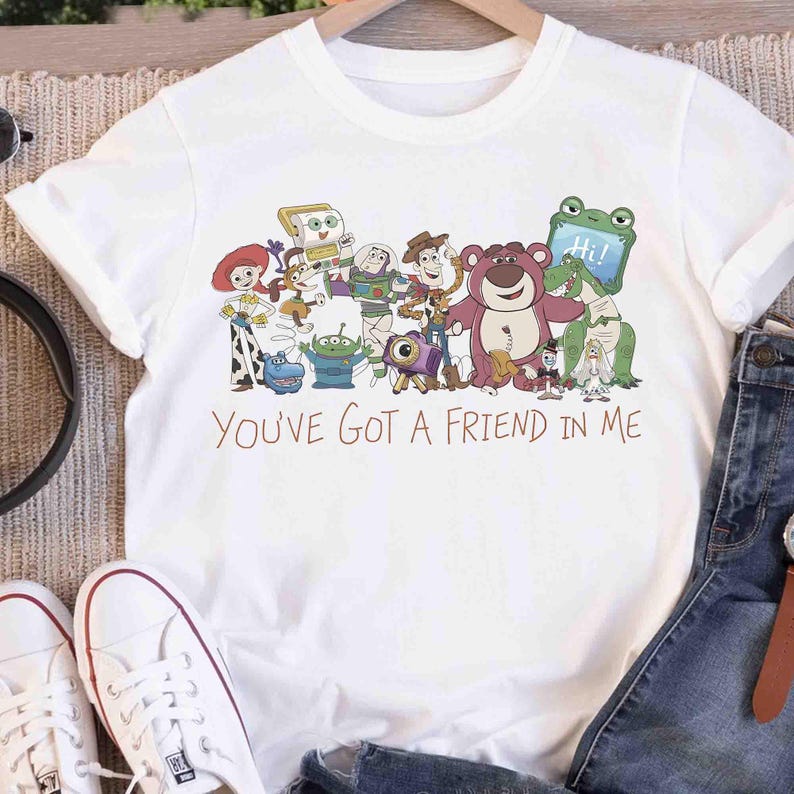 Op de afbeelding: Wit T-shirt met een kleurrijke afbeelding van Toy Story-personages, waaronder Woody en Buzz Lightyear. Het ontwerp toont meerdere personages en de tekst "You've Got a Friend in Me" in bruin.