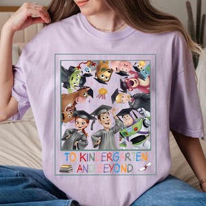 May include: A light purple t-shirt featuring a colorful graphic of Toy Story characters wearing graduation caps. The text "TO KINDERGARTEN AND BEYOND" is printed below the graphic. The shirt is a comfortable, casual style.